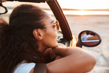Image of young thoughtful woman looking aside while riding in convertible stylish car by seaside