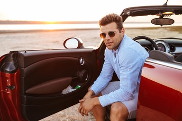 Photo of beautiful caucasian man sitting in convertible stylish car by seaside at sunrise
