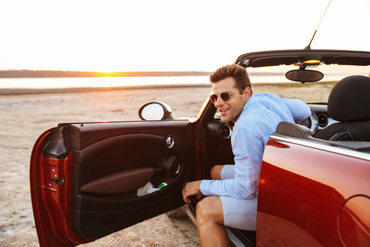Photo of handsome caucasian man getting out of convertible stylish car by seaside at sunrise