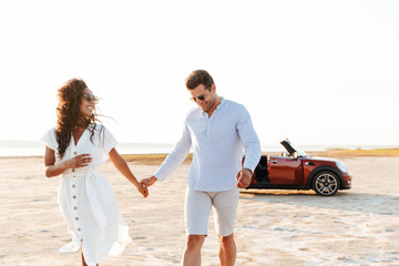 Photo of attractive multiethnic couple smiling together while walking by car on beach