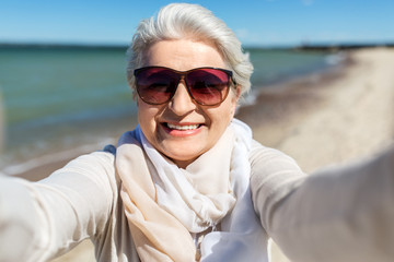 old people and leisure concept - happy smiling senior woman in sunglasses taking selfie on beach in estonia