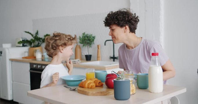 Slow motion of happy people mother and son having fun and enjoying breakfast cereal with milk in light kitchen in modern apartment. People and meals concept.