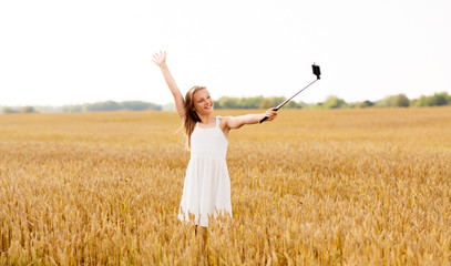 Fototapeta na wymiar technology, summer and people concept - happy young girl in white dress taking picture by smartphone on selfie stick on cereal field