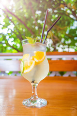 Natural citrus lemonade with slices of orange, lemon and lime in a glass on a wooden table against the background of a gazebo and bushes. Vitamin drinks concept. Bright sunshine.