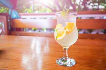 Natural citrus lemonade with slices of orange, lemon and lime in a glass on a wooden table against the background of a gazebo and bushes. Vitamin drinks concept. Bright sunshine.