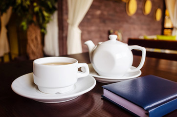 White cup of tea and teapot, notepad on a table in a cafe. The concept of tea drinking, rest, drawing up a business plan.