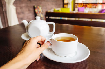 White cup of tea and teapot on a table in a cafe. Female hand holds a cup. The concept of tea drinking, relaxation.