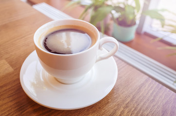 Americano coffee in a white cup on a wooden table against the background of a window, behind which trees. On a horse are plants. Cozy minimalistic still life.