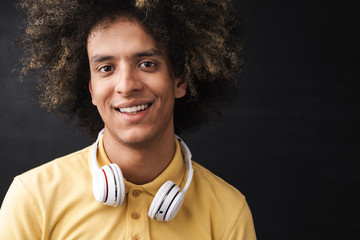 Curly young positive optimistic happy teenage guy posing over grey chalkboard wall with headphones.