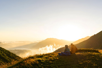 Loving couple in warm jackets sit on the top of the mountain Bzerpinsk cornice and enjoy the view of the clouds and the hacky sun. Romantic relationship. A date in the bosom of nature. © Ilona