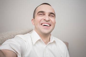 Obraz premium Attractive young man in white shirt laughs and makes a video call, sitting at home on the couch, front view, toned