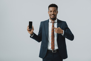 happy african american businessman pointing with finger at smartphone with blank screen isolated on grey