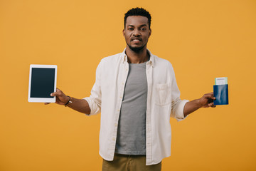 emotional african american man holding digital tablet with blank screen and passport isolated on orange