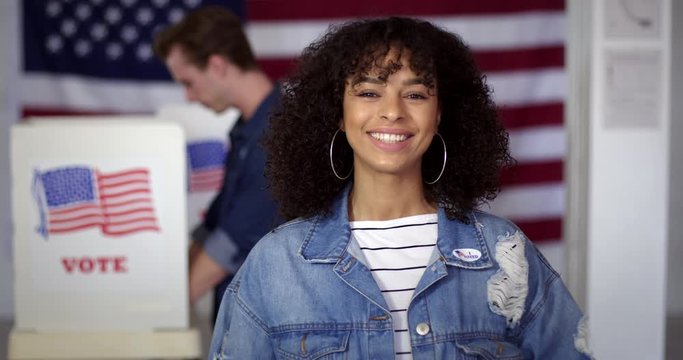 MCU Young Hispanic woman in denim jacket puts on "I Voted" sticker and smiles, standing proud in front of polling booths with US flag. Fluid head tripod, slow motion 4K 60fps