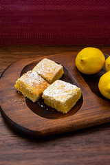 Home baked lemon squares, lemon bars, with lemons over wooden blurred background in portrait orientation 