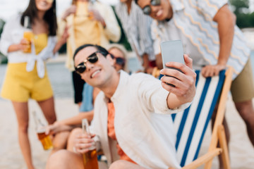 selective focus of cheerful young man taking selfie with multicultural friends having fun on beach