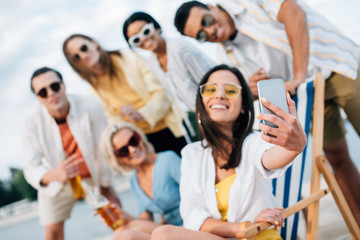 selective focus of attractive young woman taking selfie with multicultural friends having fun on beach