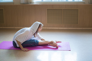 Young slim woman in the white hood sitting on the yoga mat holding the foot and bending it