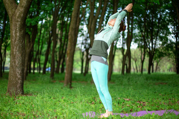 Thin brunette girl plays sports and performs beautiful and sophisticated yoga poses in a summer park. Green lush forest on the background. Woman doing exercises on a yoga mat