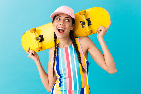 Image of cute joyful woman wearing cap looking aside at copyspace while carrying yellow skateboard on her shoulders