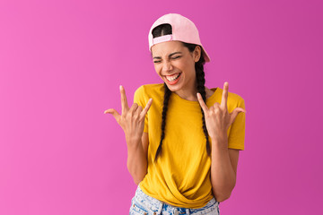 Photo of young joyful woman wearing cap backwards smiling and showing rock sign