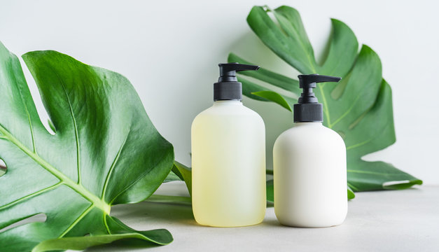 Cosmetic set of two blank label bottles for mockup packaging of skincare product cream, shampoo, conditioner on grey background with green leaves. Natural beauty product concept.