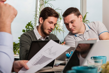 Obraz premium two businessmen discussing price calculation during a meeting. they sit at the table with open laptops holding paper sheets