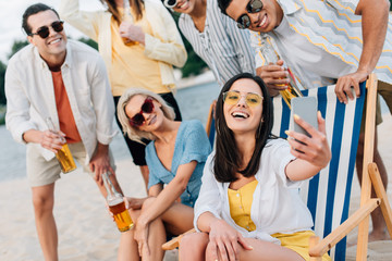 attractive cheerful woman taking selfie with multicultural friends having fun on beach