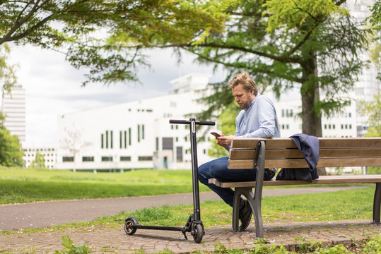 Businessman with E-Scooter sitting on bench in city park using smartphone, Essen, Germany