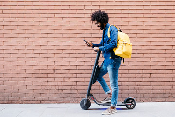 Young man with backpack and E-Scooter standing in front of brick wall looking at cell phone