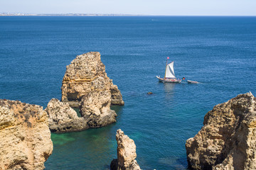 Large sailboat with a small boat in tow passing by the colourful  cliffs of  Ponta da Piedade in Algarve. Photo taken from above. Rocks and turquoise sea water in foreground, town of Lagos in distance