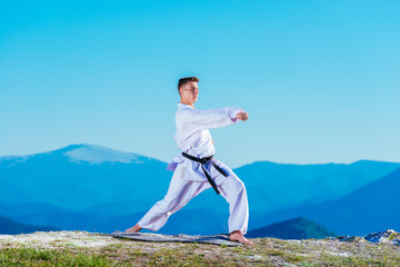 Karate man in a kimono performs a front hand kick (Choku-zuki) while standing on the green grass on top of a mountain.