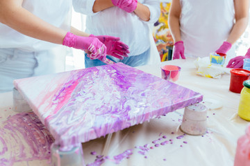 Woman painter holding hair dryer while female beginnres studying creating fluid acrylic abstract painting in art therapy class, dropping paints on canvas. Teamwork, study, art therapy concept.