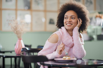 Portrait of charming positive mixed race woman in pink turtleneck sweater sitting in confectionery and eating gateau.
