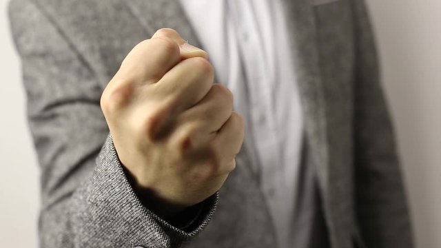 Solid caucasian man in gray jacket and speckled shirt on white background pulling fist up. close-up on male fist shaking in front of camera. threat gesture, concept of aggression, warning, punishment