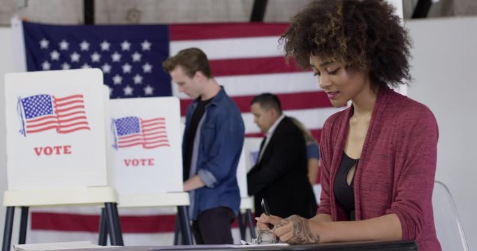 MS Young mixed-race woman staffing desk at polling station with various voters in background, She looks up and smiles at camera. US flag on wall. Hand-held, real time 4K