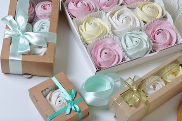 Marshmallows of different colors and shapes. Some in the shape of a flower. Stacked in gift boxes. Nearby are colored ribbons for dressing boxes.