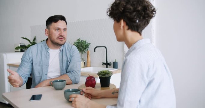 Aggressive guy husband is yelling at unhappy woman in kitchen fighting gesturing sitting at table together shouting at each other. Conflict, problems and family concept.