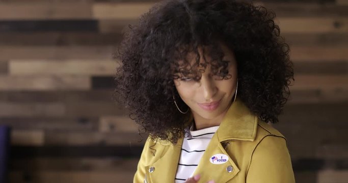 MCU Young Hispanic woman in yellow leather jacket puts on "I Voted" sticker and smiles. Wooden plank background. Slow motion hand-held 4K 60fps