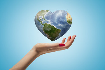 Side closeup of woman's hand facing up and levitating small heart-shaped Earth on light blue gradient background.