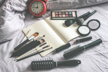Make up cosmetic and brushes, alarm clock and blank page notepad on the blanket on the bed background. Female morning makeup tips mockup.