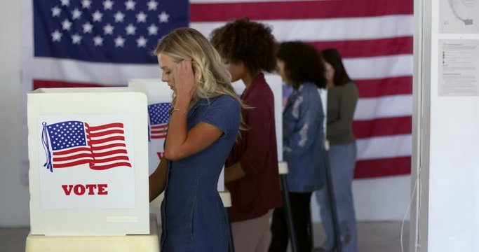 MS Four women voters of various demographics filling in ballots at voting booths in polling station with US flag. Locked off continuous shot, slow motion 4K