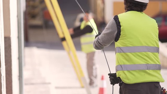 Slow motion shot of a technician worker pulling down an optical fiber cable from a roof of a building with another technician blurred in the background