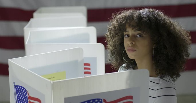 MS Young Hispanic woman in polling station, voting in a booth with US flag in background. Serious expression from high viewpoint. Slow motion 4K 60fps