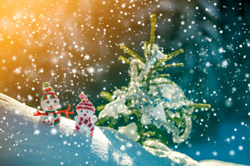 Two small funny toys baby snowman in knitted hats and scarves in deep snow outdoors near pine tree branch. Happy New Year and Merry Christmas greeting card.