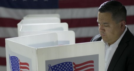 MCU crane move from side following Hispanic man, entering booth to cast vote at polling station. US flag in background. Slow motion 4K 60fps - Powered by Adobe