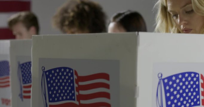 CU crane move up from booths to young blonde woman casting vote with others at polling station. She looks up to think and looks down again. Slow motion 4K 60fps