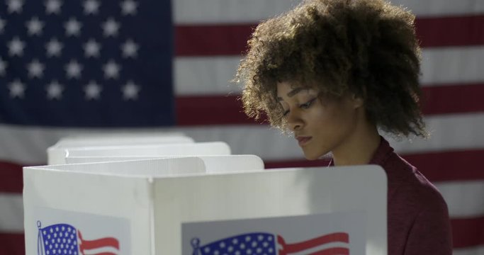 MCU crane move from side to young mixed race woman, alone casting vote at polling station. She looks across and smiles. US flag in background. Slow motion 4K 60fps