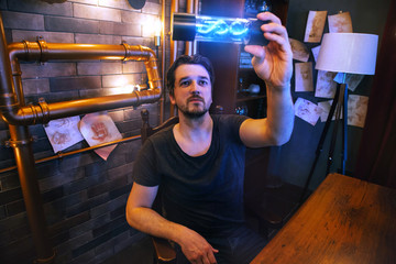 Portrait of a young man in a steampunk scientist’s office. The doctor holds in his hands a flask with a dna structure that glows in blue. Handsome bearded intelligence spy guy.
