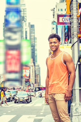 Obraz na płótnie Canvas Young Happy Mix Race American Man traveling in New York City in hot summer, wearing orange tank top, beige pants, standing on intersection of street in Times Square of Manhattan, looking, smiling..